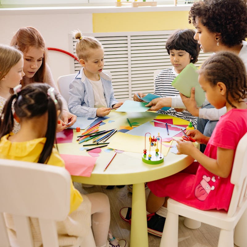 teacher sitting with kids at their class table. They are doing art.